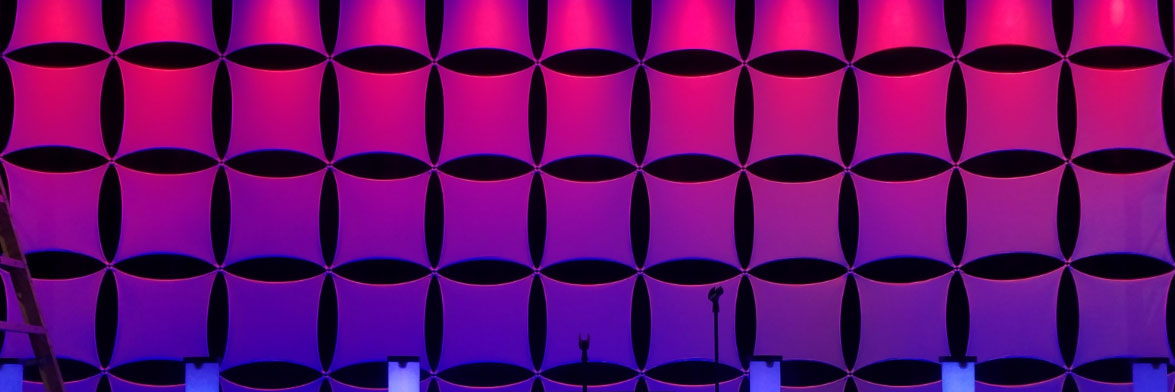 A Stretch Geometric Fabric Panel Wall Illuminated With Purple And Pink Lighting